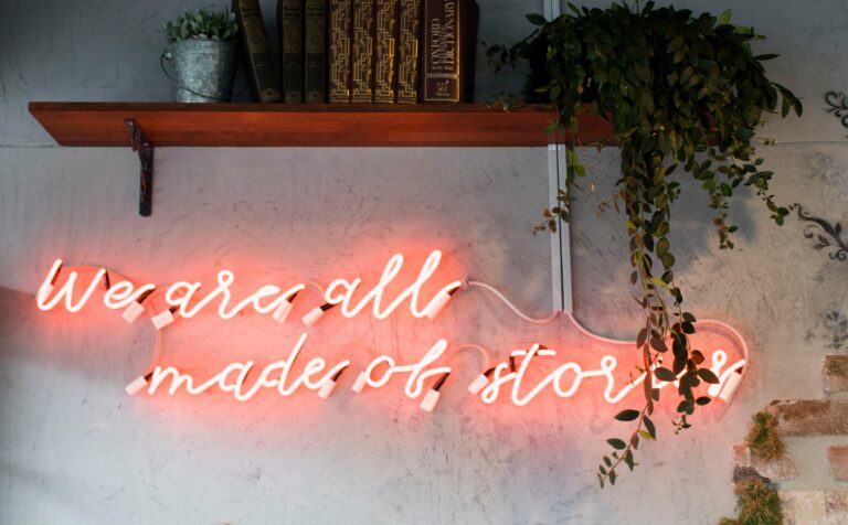 Bookshelf with books and plants on a wall above a red neon sign that says "We are all made of stories" Image by socialcut-FluPNkHfCTs