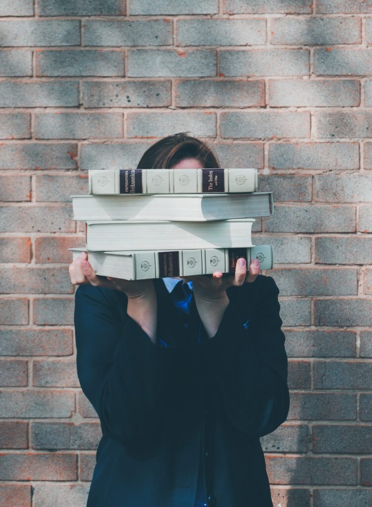 A person stands in front of a grey brick wall, holding a stack of books in front of their face. Image by Siora Photography.